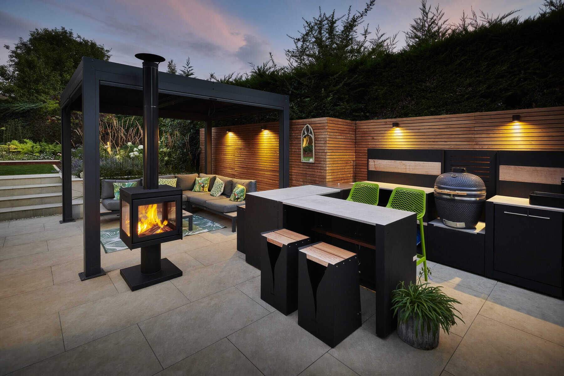 Covered outdoor seating area with a wood-burning stove and modern outdoor kitchen on a paved patio at dusk