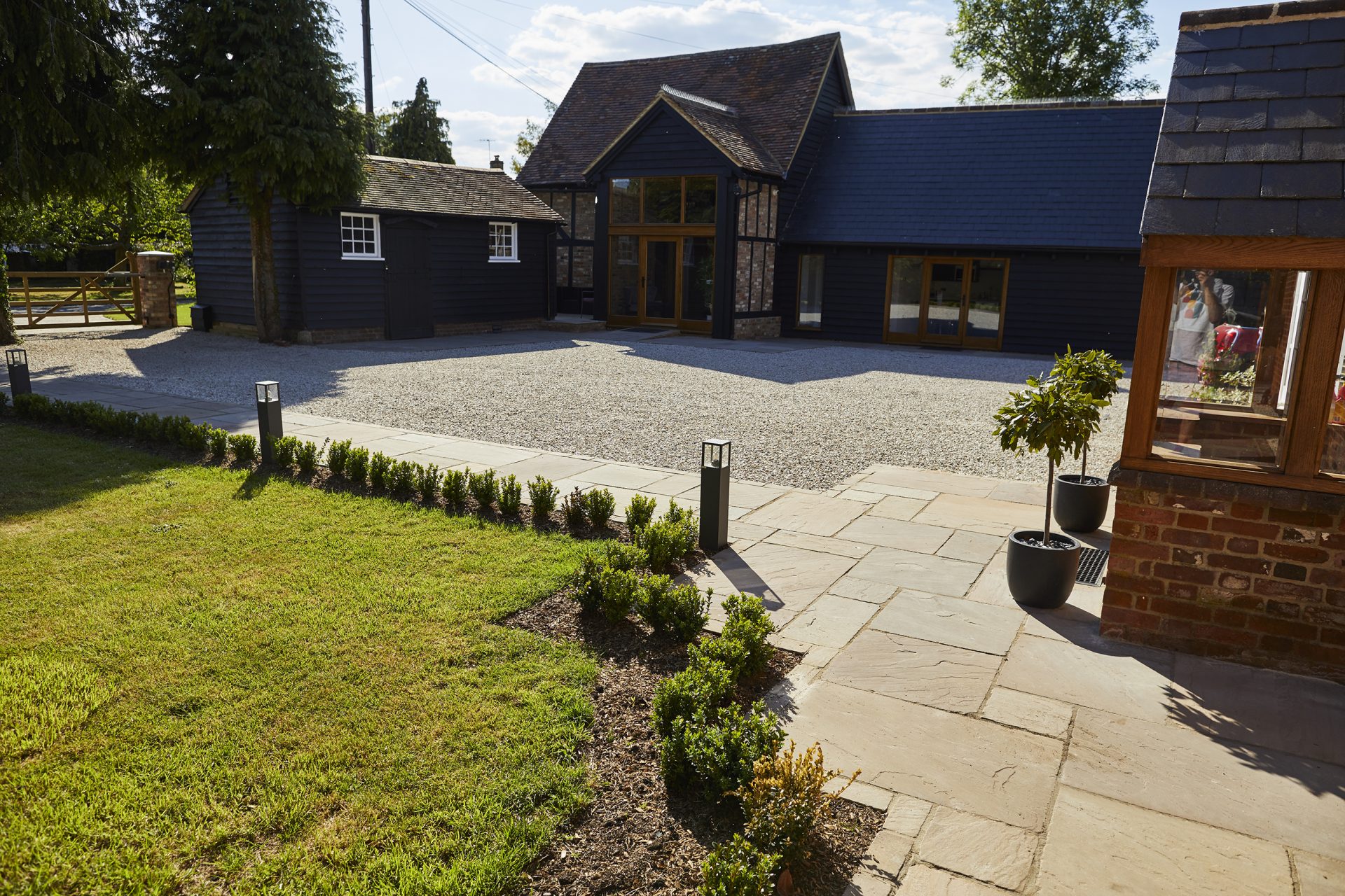 Gravel courtyard and stone-paved path beside a black timber-clad home, with a lawn, low hedging and modern bollard lights.