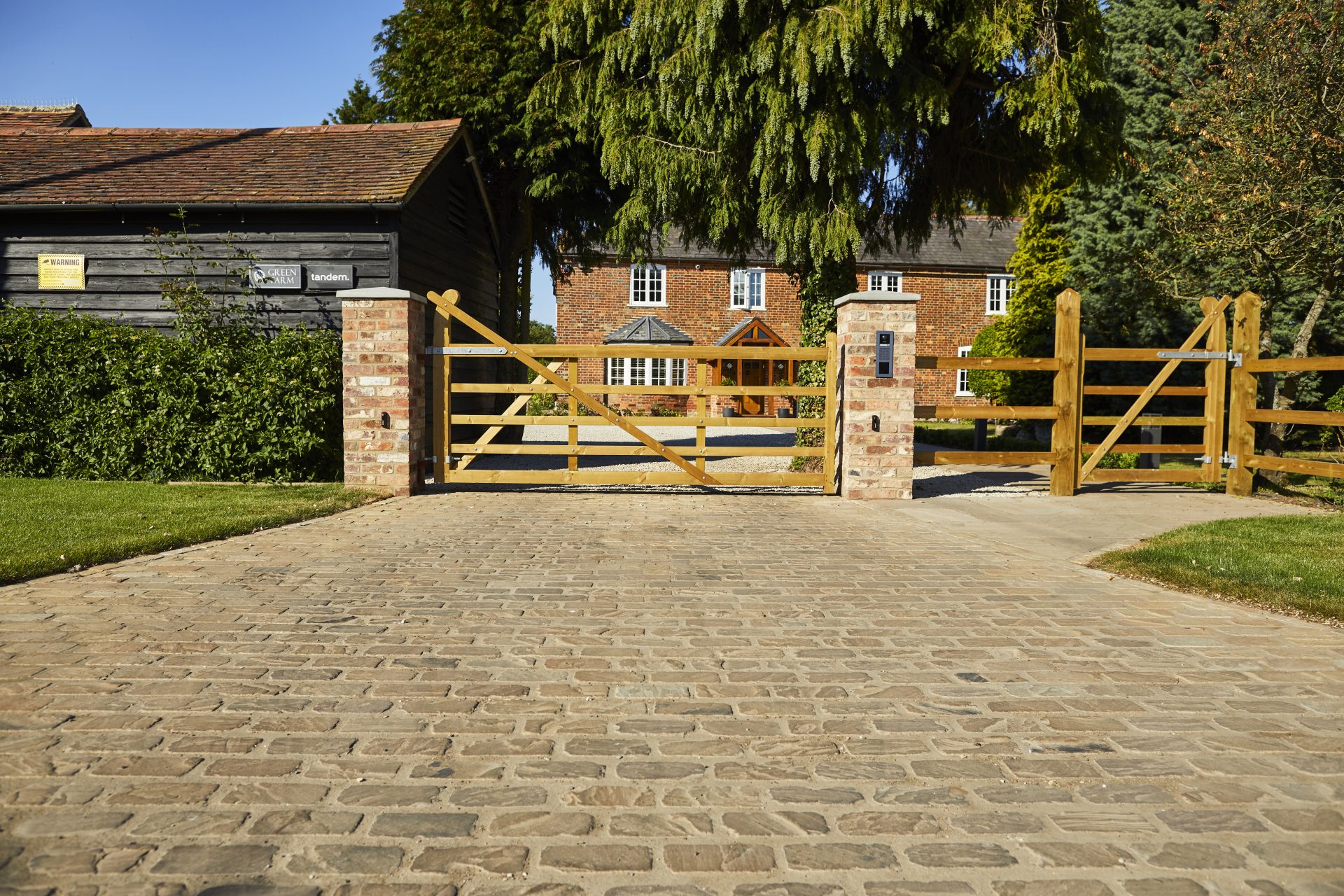 Cobblestone driveway leading to timber entrance gates with brick pillars in front of a large country house.