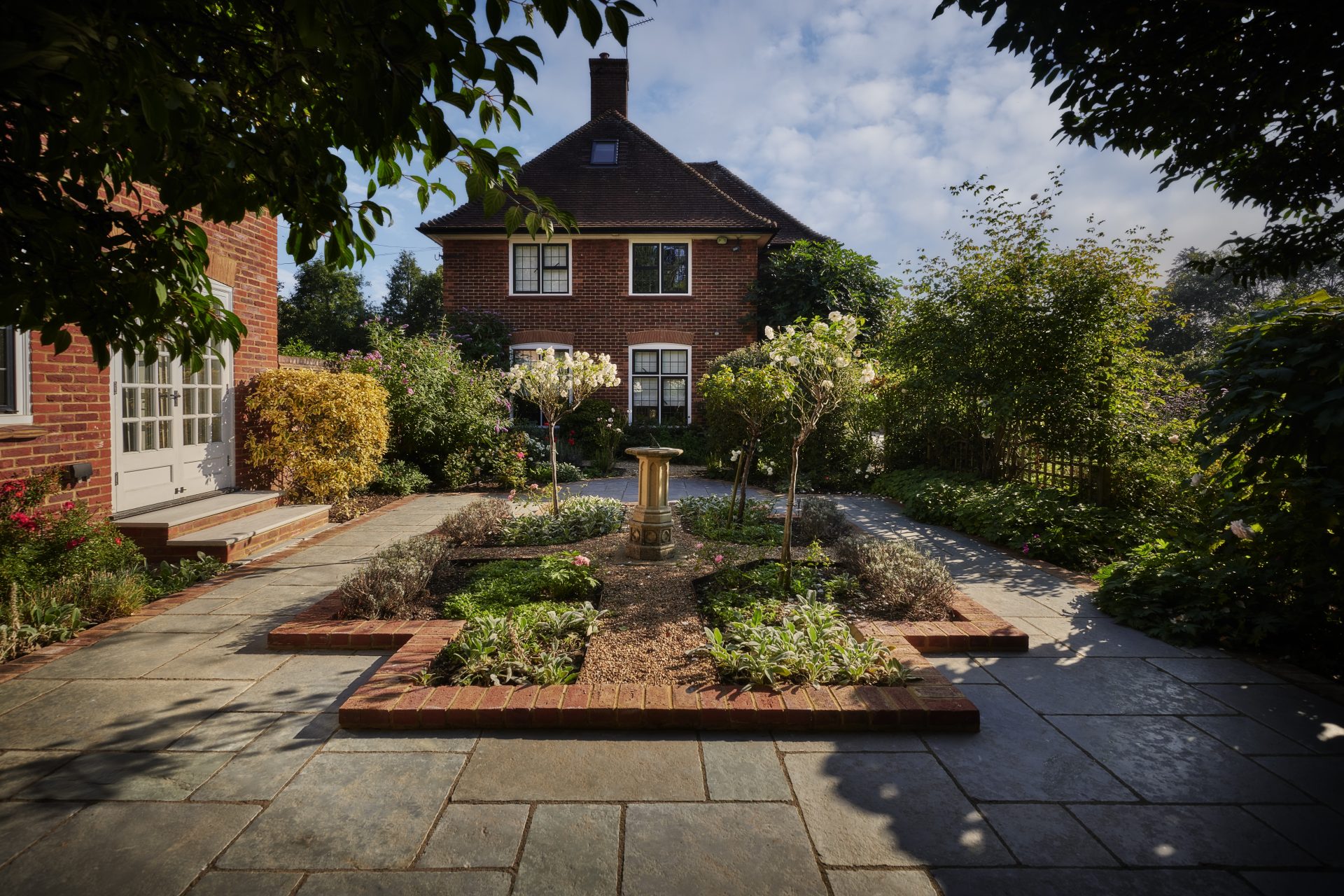 Formal garden courtyard with stone paving, brick-edged planting beds, ornamental trees and a central pedestal feature.