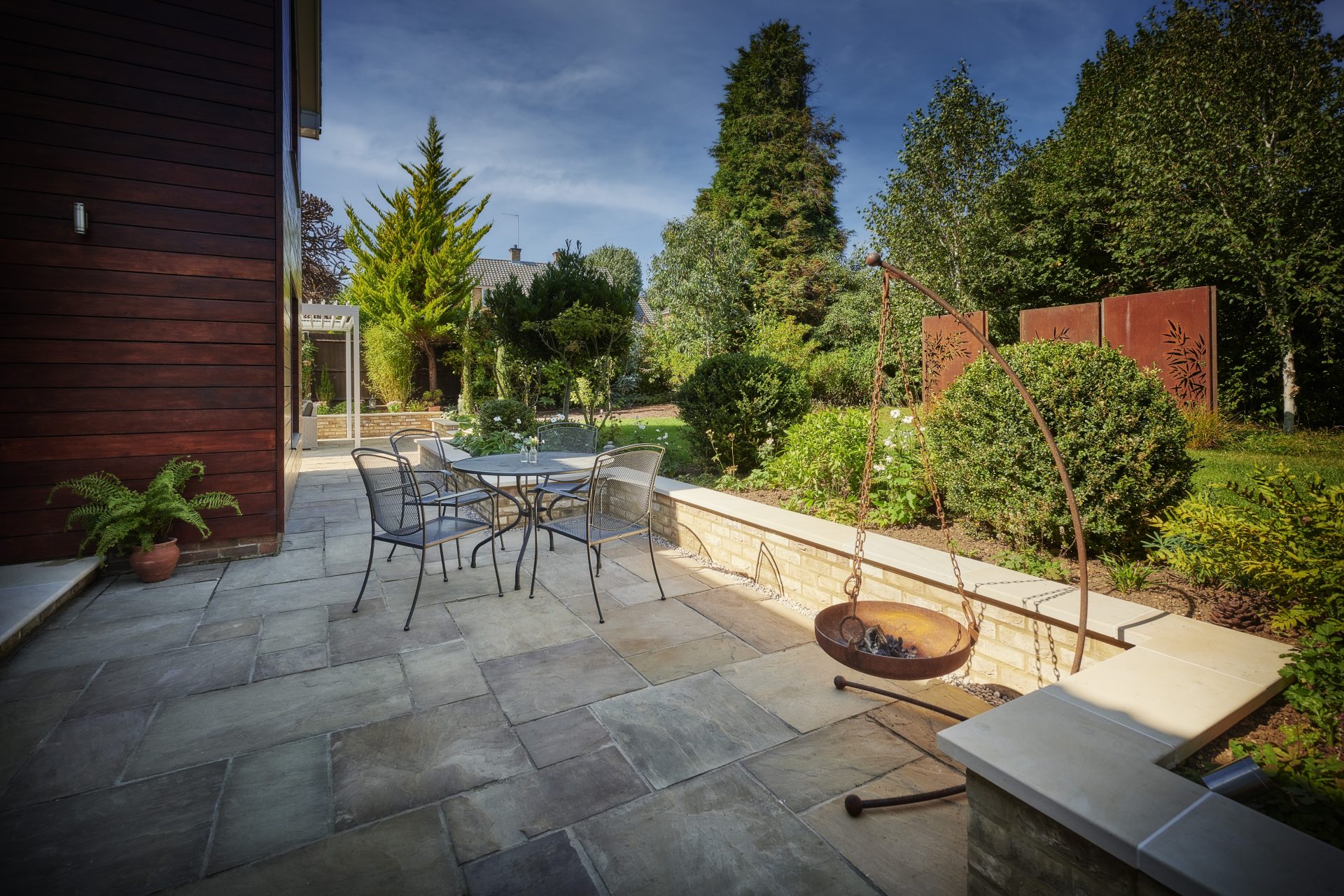Stone-paved patio with metal dining furniture, a suspended fire bowl, raised planting beds and decorative weathered-steel screens.