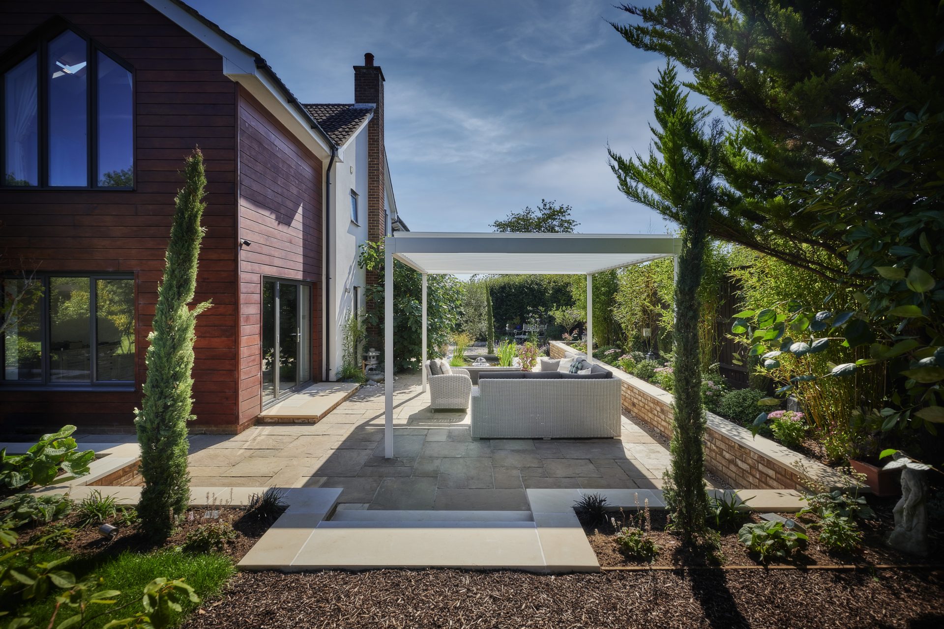 Modern garden terrace with a white pergola, outdoor lounge seating, stone paving and raised planted borders beside a timber-clad house.