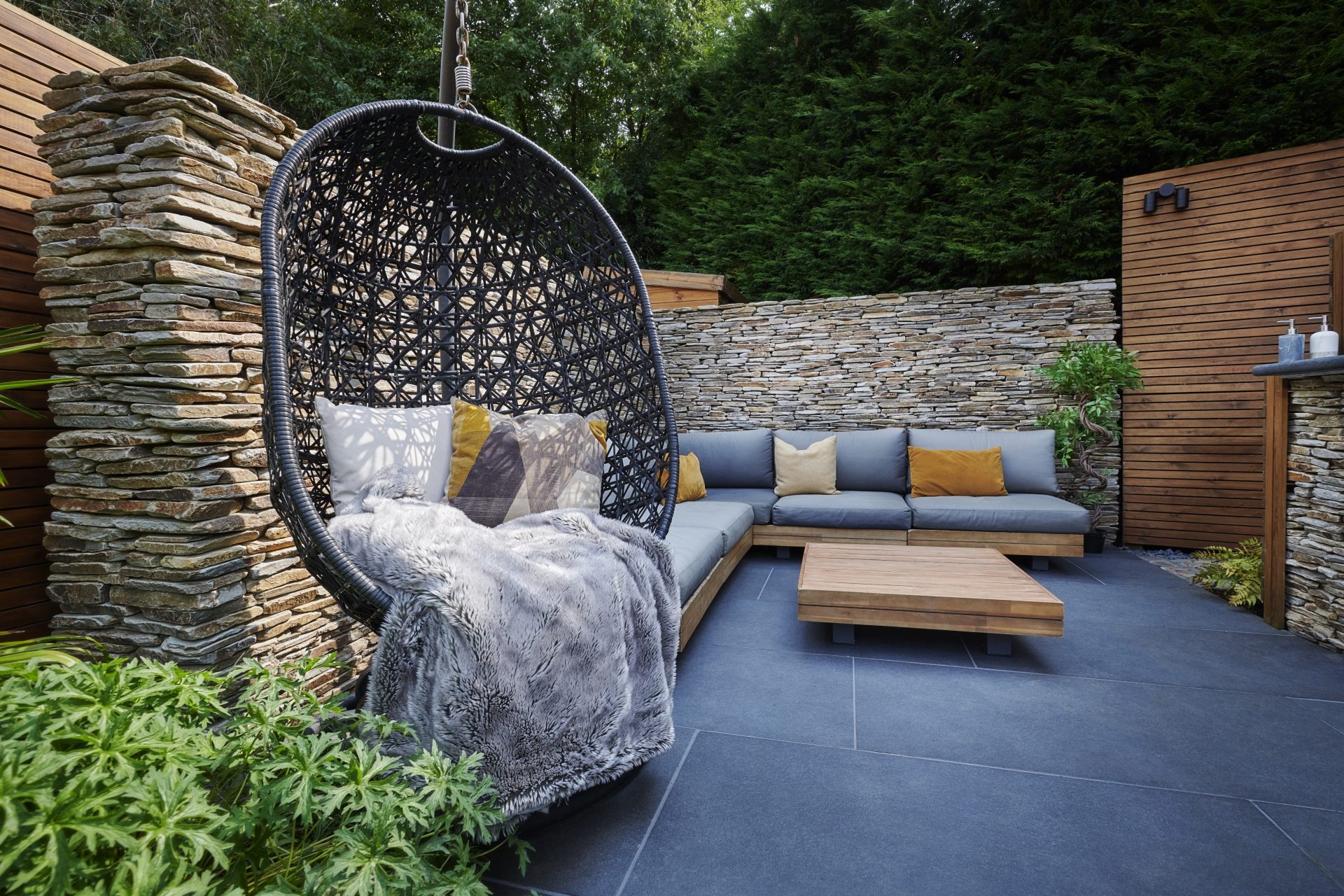 Contemporary garden seating area with a hanging egg chair, grey outdoor sofa, dark porcelain paving and stacked-stone walls.