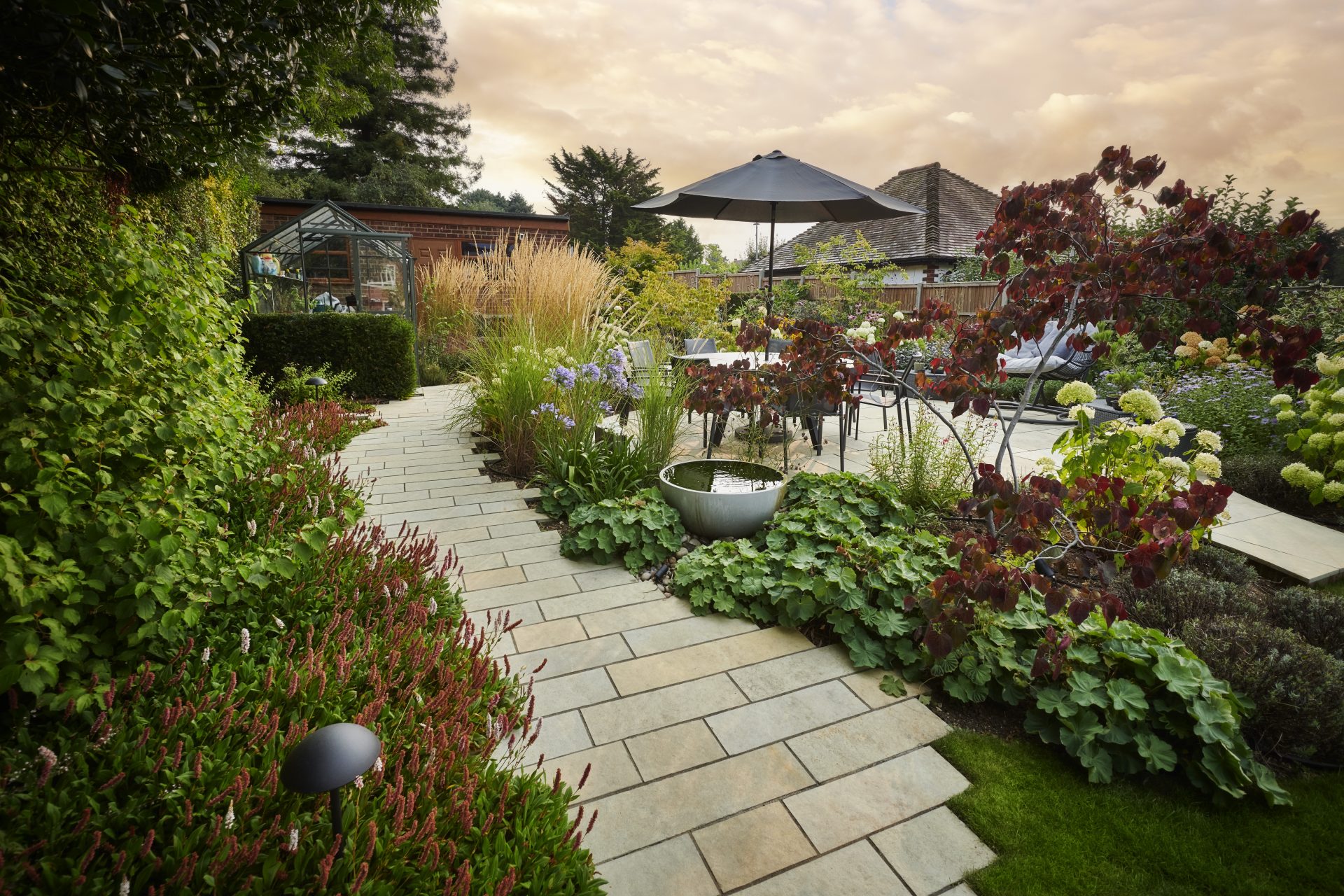 Landscaped garden path leading to a paved dining terrace, bordered by ornamental grasses, flowering plants and a small water bowl.