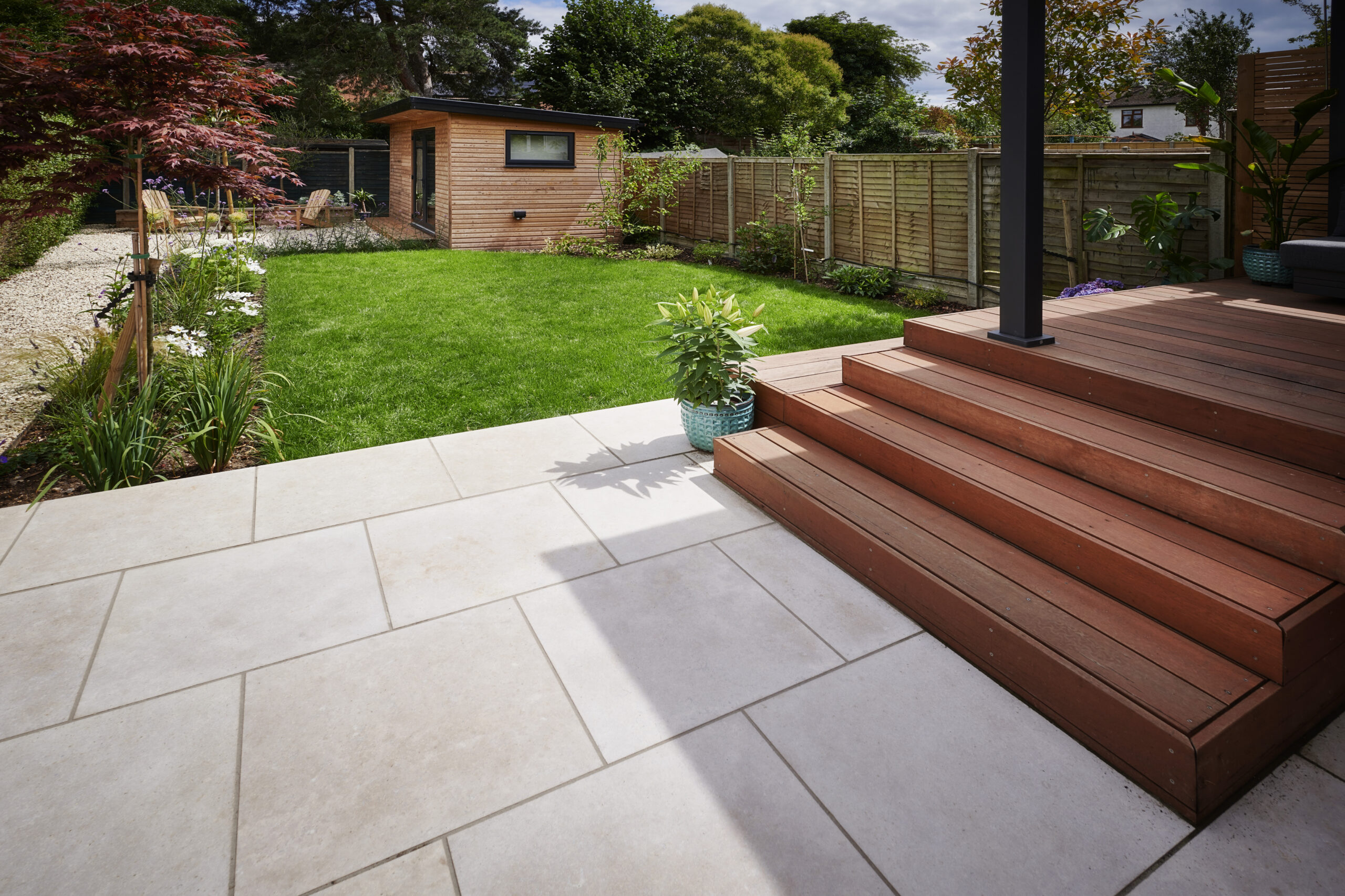Large porcelain paved patio with timber deck steps, lawn, planting and a garden room.
