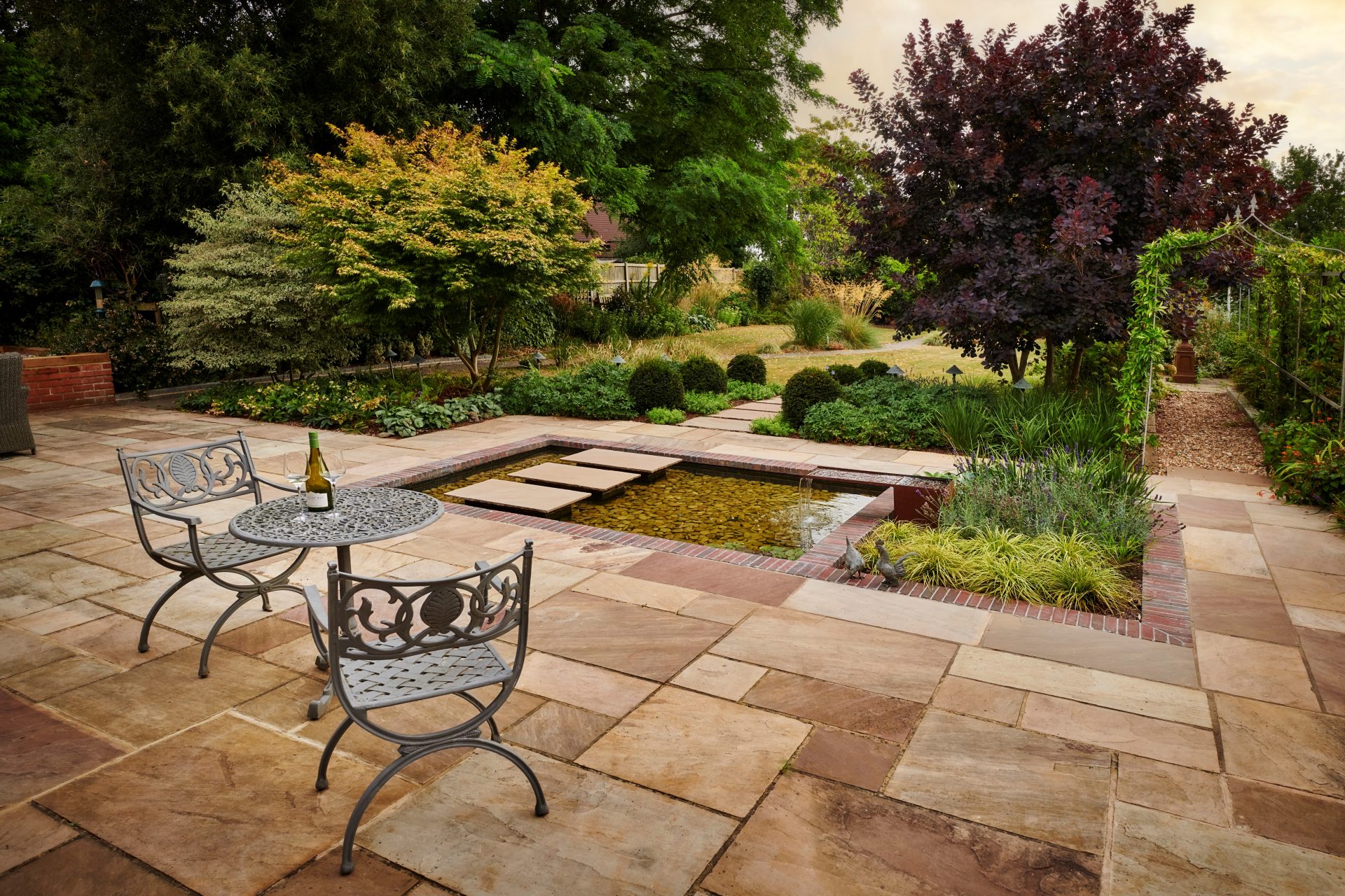Sandstone patio with metal dining furniture beside a rectangular pond, stepping stones and mature planted borders.